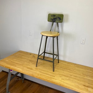 May include: A green metal industrial stool with a wooden seat and a backrest. The stool is sitting on a wooden table.