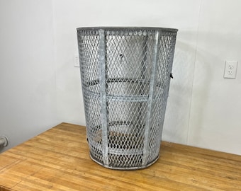 May include: A tall, cylindrical metal trash can with a diamond-patterned mesh design. The can is silver and has a sturdy, industrial appearance. It sits on a light-colored wooden surface.