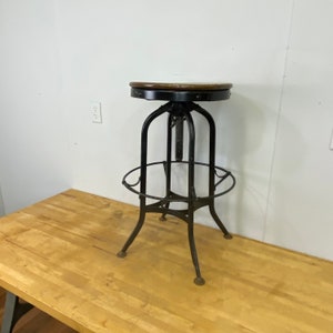 May include: Black metal adjustable height bar stool with a wooden seat. The stool has a footrest and a ring around the base.