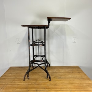 May include: A black metal industrial style table with a wooden top and a smaller shelf. The table has a distressed finish and is on wheels.