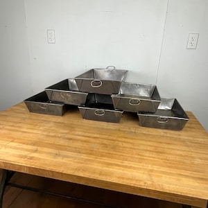 May include: Six silver metal trays stacked in a pyramid shape on a wooden table. The trays have a simple design with a handle on the top and a lip around the edge.