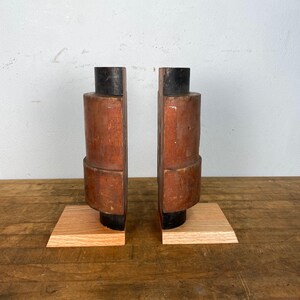 May include: A pair of wooden bookends with a dark brown finish and a square base. The bookends are made of two pieces of wood that are joined together to form a cylinder shape. The bookends are a unique and decorative way to organize books.