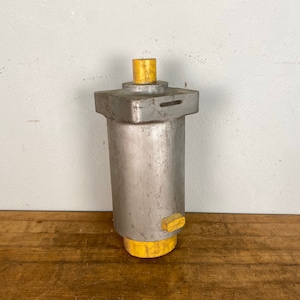 May include: A silver metal cylinder with yellow wooden caps on the top and bottom. The cylinder has a small rectangular opening on the side.