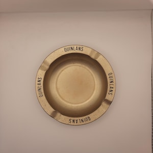 May include: A vintage, circular ashtray with a gold finish. The word "QUINLANS" is printed in black around the rim. The ashtray has a shallow bowl and three small rests for cigarettes.