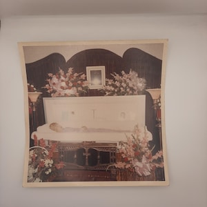 May include: A framed photograph of a funeral scene. A person lies in an open casket, surrounded by floral arrangements in shades of pink, red, and white. The casket is set on a dark wooden stand.