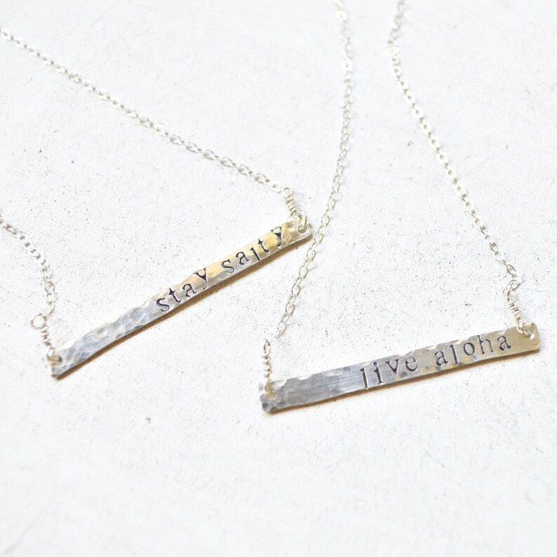 May include: Two silver necklaces with hammered metal bar pendants. The top pendant says "stay salty" and the bottom pendant says "live aloha".