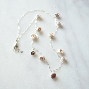 Peut inclure: Un collier en chaîne argentée avec plusieurs perles en forme de coquille de couleur blanche et brune. Le collier a une fermeture à fermoir.