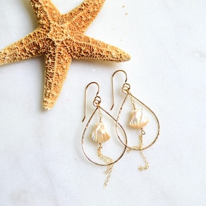 Puede incluir: Pendientes dorados en forma de lágrima con un dije de concha y una cadena colgante. Los pendientes se muestran con una estrella de mar sobre una superficie blanca, evocando un tema de playa. Un accesorio delicado.
