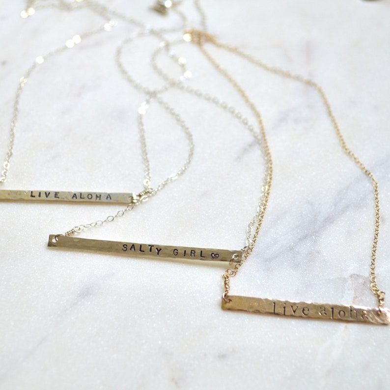 May include: Three silver necklaces with hammered bar pendants. The pendants are engraved with the phrases "Live Aloha", "Salty Girl", and "Live Aloha".