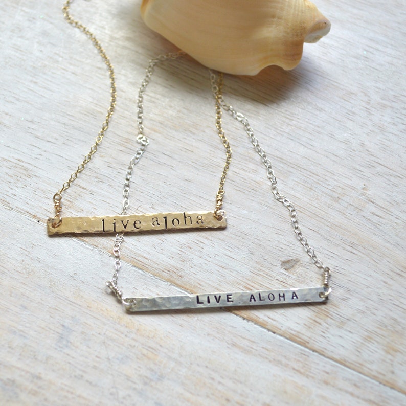 May include: Two gold necklaces with hammered metal bar pendants. The top necklace has the text "live aloha" on the pendant. The bottom necklace has the text "LIVE ALOHA" on the pendant.