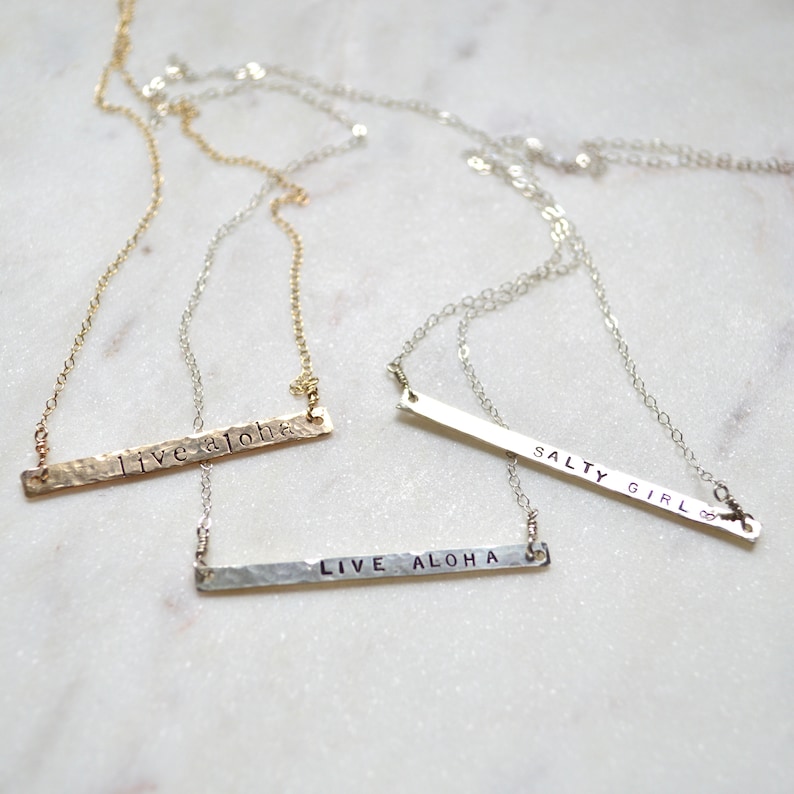 May include: Three gold and silver necklaces with hammered bar pendants. The pendants are engraved with the phrases "Live Aloha", "Live Aloha", and "Salty Girl".