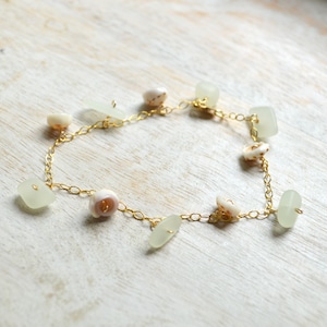 Peut inclure: Un collier en chaîne dorée avec des breloques en verre de mer blanc et en coquillage. Le collier est sur une surface en bois blanc.