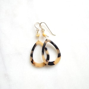 May include: A pair of gold teardrop earrings with a black and white tortoiseshell pattern. The earrings have a small gold bead at the top.