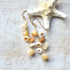 Peut inclure: Boucles d'oreilles pendantes dorées ornées de petits coquillages et de pierres précieuses jaune-orangé. Les boucles d'oreilles sont suspendues à des crochets et des chaînes dorées. Une étoile de mer blanche est en arrière-plan.