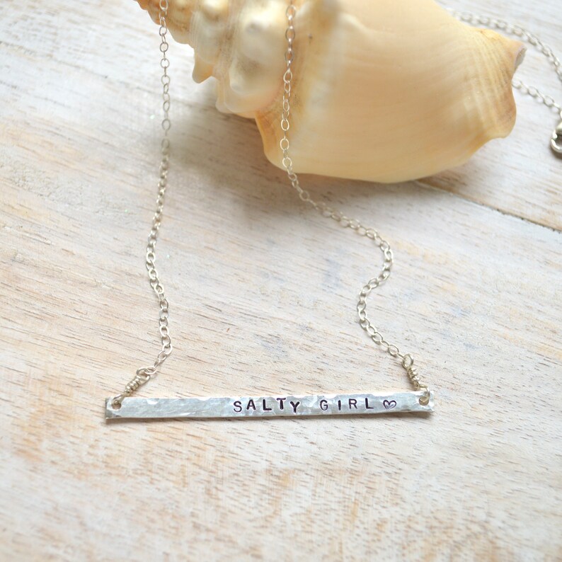 May include: A silver necklace with a hammered bar pendant that says "Salty Girl" with a heart symbol. The necklace is on a white wooden surface.