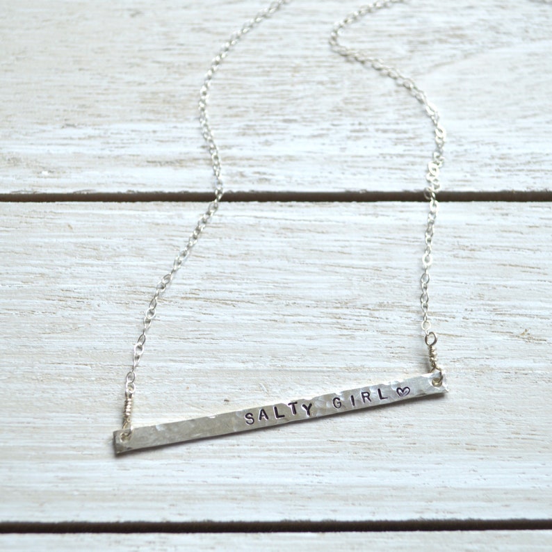 May include: A silver necklace with a hammered bar pendant that says "Salty Girl" with a heart symbol.