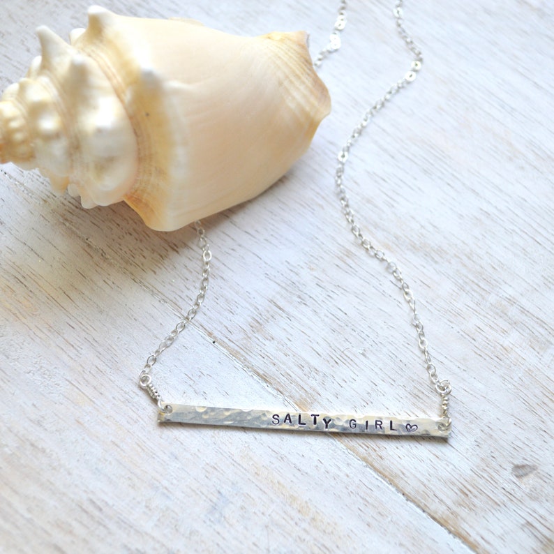 May include: Silver necklace with a hammered bar pendant that says "Salty Girl" with a heart symbol. The necklace is displayed on a white wooden surface with a seashell.