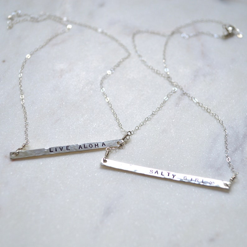 May include: Two silver necklaces with hammered bar pendants. The top pendant says "LIVE ALOHA" and the bottom pendant says "SALTY GIRL".