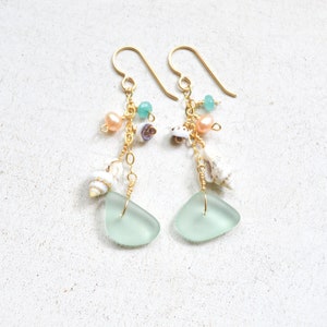 Op de afbeelding: Een paar oorhangers met een lichtblauwe glazen traanvorm, een gouden ketting en een verscheidenheid aan kleine witte schelpen, roze en groene kralen.