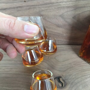 May include: Four clear glass shot glasses filled with amber liquid. The glasses are stacked on top of each other, with the top glass being held by a hand.