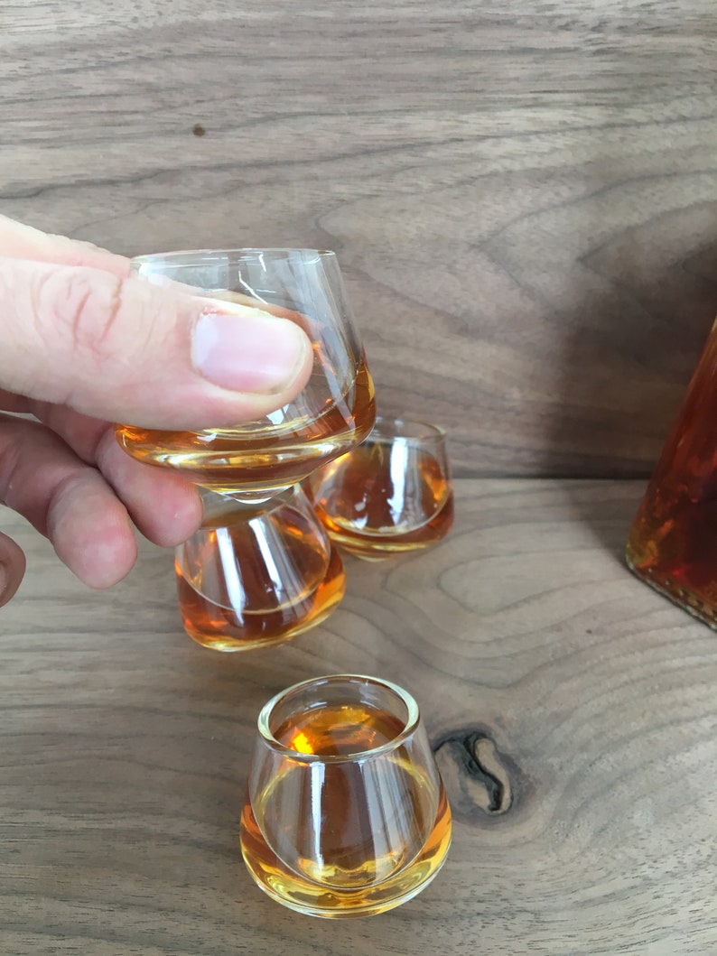 May include: Four clear glass shot glasses filled with amber liquid. The glasses are stacked on top of each other, with the top glass being held by a hand.