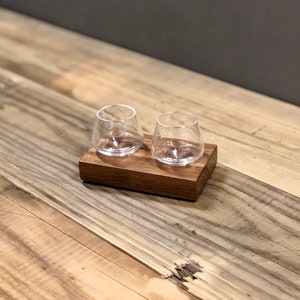 May include: Two clear glass shot glasses on a wooden coaster. The coaster is made of dark wood and has a rectangular shape.