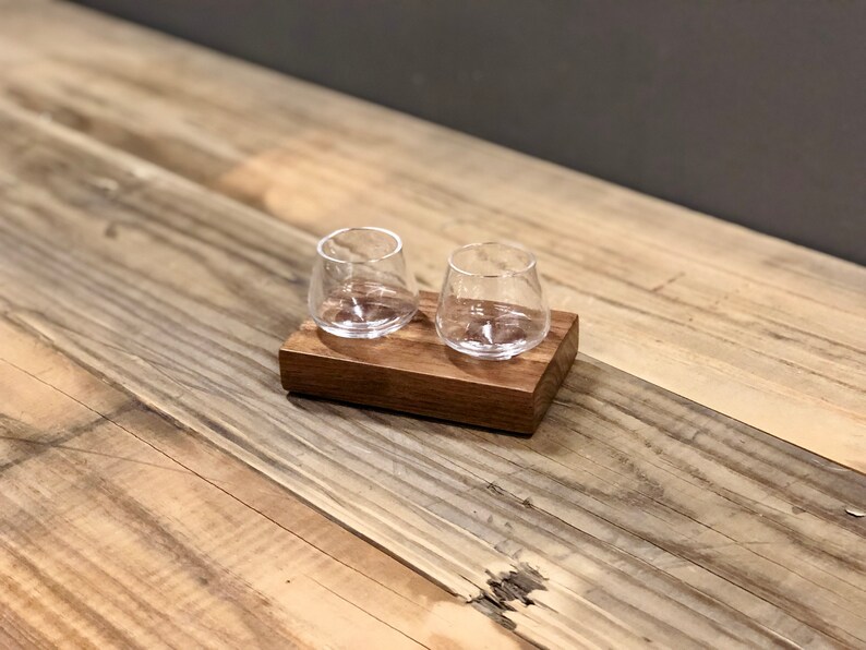 May include: Two clear glass shot glasses on a wooden coaster. The coaster is made of dark wood and has a rectangular shape.
