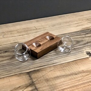 May include: A wooden tray with two indentations for holding small glasses. The tray is made of dark wood and has a natural finish. Two clear glass tumblers are sitting on the tray.