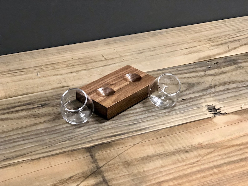 May include: A wooden tray with two indentations for holding small glasses. The tray is made of dark wood and has a natural finish. Two clear glass tumblers are sitting on the tray.