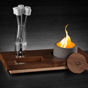 May include: A wooden tray with a built-in fire pit and a small, round, wooden chopping board. The fire pit is lit and has a small, orange flame. There are four white marshmallows on sticks in a glass vase.