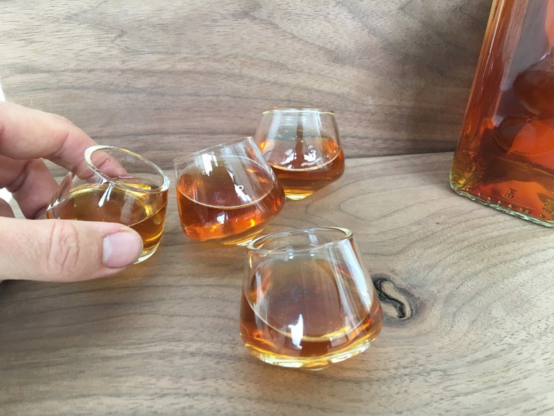 May include: Four clear glass shot glasses filled with amber liquid, sitting on a wooden surface.