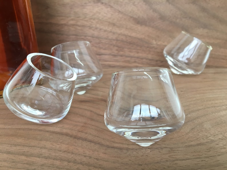 May include: Four clear glass shot glasses with a unique, rounded, and slightly tilted design. The glasses are arranged on a wooden surface.
