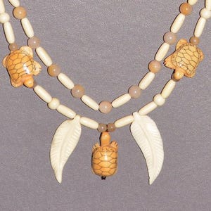 May include: A two-strand necklace featuring turtle and feather pendants. The necklace has cream-colored beads and turtle pendants with a light brown shell design. The necklace also has light brown and cream-colored feather pendants.