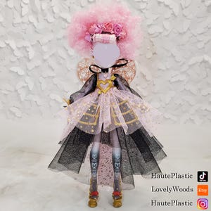 May include: A doll with a pink afro wig, a floral headband, and a heart-shaped pendant. The doll is wearing a pink and black dress with gold accents and gold shoes. The doll's legs are patterned with a floral design. The background is white.