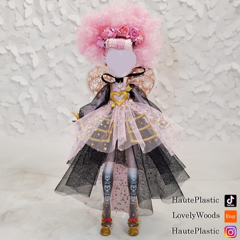 May include: A doll with a pink afro wig, a floral headband, and a heart-shaped pendant. The doll is wearing a pink and black dress with gold accents and gold shoes. The doll's legs are patterned with a floral design. The background is white.