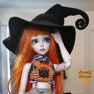 May include: A doll with long orange hair wearing a black witch hat with a curled tip. The doll is also wearing an orange crop top with a black cat design and a striped scarf. The doll is a Halloween-themed item.