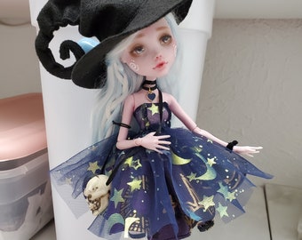 Puede incluir: Muñeca vestida de bruja, con sombrero negro, vestido azul oscuro con estrellas y lunas doradas, y calcetines a rayas. La muñeca tiene piel pálida, pelo azul y un pequeño bolso con forma de calavera.