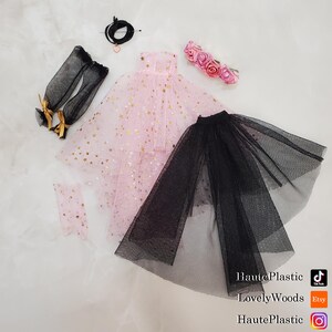 May include: A collection of doll clothing and accessories, including a pink dress with gold star accents, a black tulle skirt, black arm warmers with gold bows, and a pink floral headband. A black necklace with a heart pendant is also included.