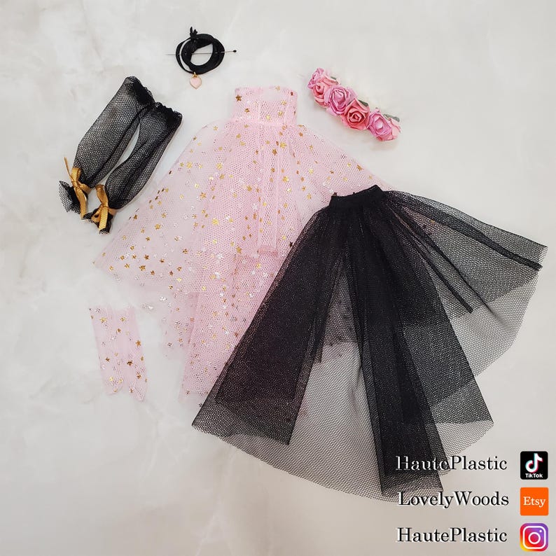 May include: A collection of doll clothing and accessories, including a pink dress with gold star accents, a black tulle skirt, black arm warmers with gold bows, and a pink floral headband. A black necklace with a heart pendant is also included.
