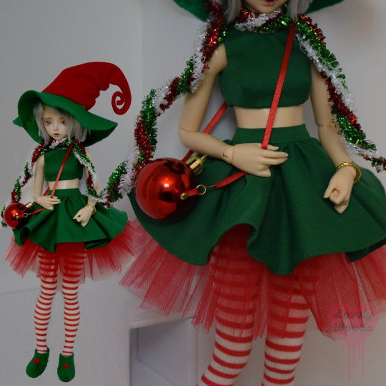 May include: Two dolls dressed in Christmas elf costumes. The outfits include green crop tops and skirts, red and white striped leggings, and red tulle underskirts. Each doll wears a red and green elf hat and carries a red ornament purse. Tinsel accents the outfits.
