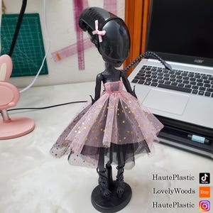 May include: A black doll with a large head and a pink bow, wearing a pink and gold star-patterned dress. The doll has black boots and is standing on a black base. The doll is next to a laptop.