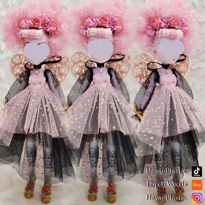 May include: Three dolls with pink afro wigs adorned with roses. They wear pink dresses with gold star accents, black tulle skirts, and gold shoes. Each doll has a heart-shaped face and decorative wings.