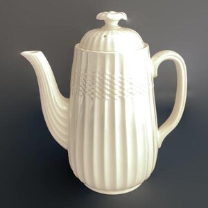 May include: Cream-colored ceramic teapot with a lid and handle. The teapot features vertical ribbed detailing on the body and a woven pattern around the middle. The spout is curved, and the handle is arched. The lid has a small floral-shaped knob.