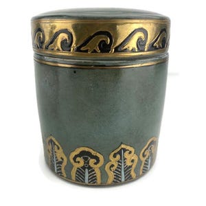 May include: A cylindrical ceramic jar with a lid, featuring a muted sage green base color. The lid is decorated with a gold and black wave pattern, while the base of the jar is adorned with a repeating gold and black leaf-like motif.