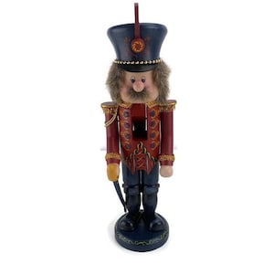 May include: A wooden nutcracker figurine in a soldier's uniform. The figurine has a dark blue hat with gold accents, a red jacket with gold trim, and black boots. It is standing on a dark blue base.