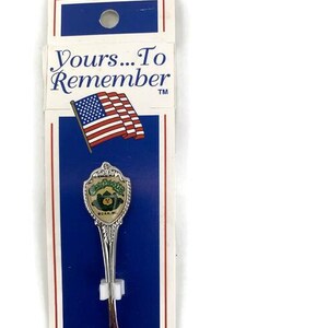 Peut inclure: Une cuillère souvenir en argent avec un motif vert et or représentant un ours et les mots "Great Smoky Mountains National Park". La cuillère est emballée sur une carte bleue et blanche avec les mots "Yours...To Remember" et un petit drapeau américain.