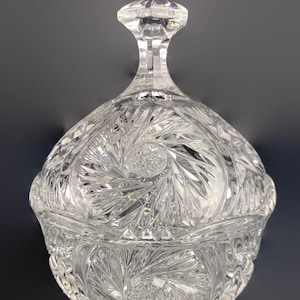 May include: A clear, cut-glass candy dish with a lid and a decorative handle. The dish features intricate starburst and geometric patterns. The clear glass reflects light, creating a sparkling effect. The dish is designed for serving candies or small treats.