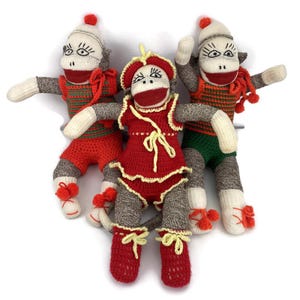 May include: Three handmade sock monkey dolls. The monkeys have grey bodies, white hands and feet, and red and white faces. They wear red and green outfits with pom-pom accents. One monkey is dressed in a red dress with yellow trim.