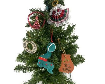 May include: A small artificial Christmas tree decorated with various ornaments. Ornaments include a beaded angel, a red and green plaid skirt, a green worm-like figure, and a tag that says "Deck them halls y'all!"