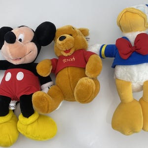May include: Three plush Disney character toys: Mickey Mouse in red shorts, Winnie the Pooh in a red shirt, and Donald Duck in a blue sailor suit with a red bow tie. The toys are soft and colorful.